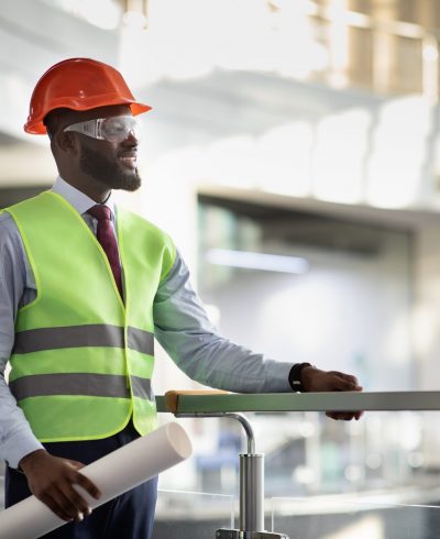 Smiling african american young man in protective vest, helmet and glasses architect visiting building site, holding rolled drawing, looking at copy space, side view, panorama